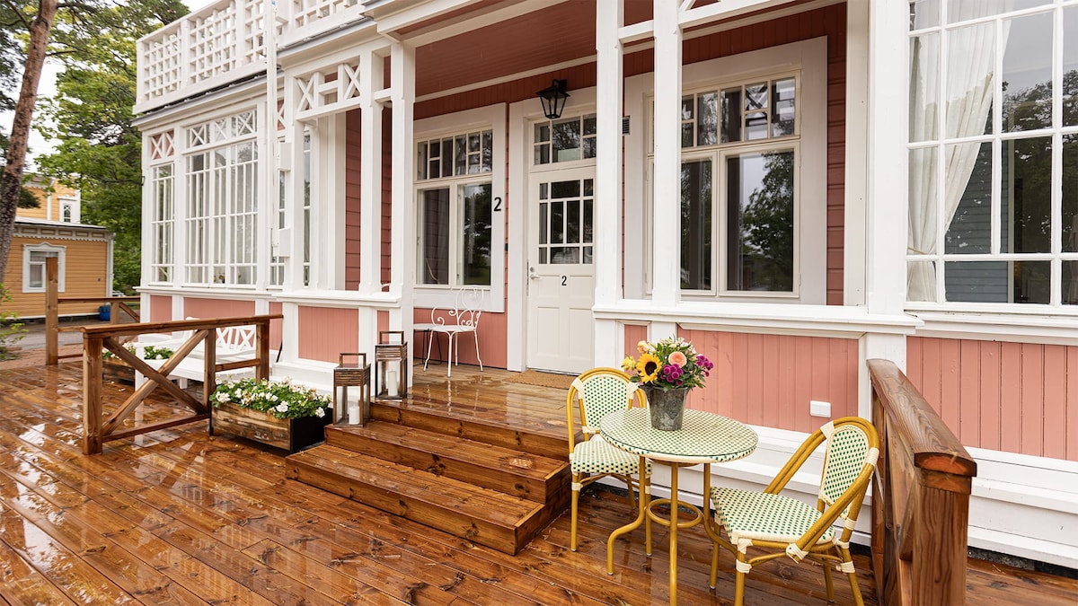A welcoming entrance features wooden steps leading to a charming terrace adorned with potted flowers. Two stylish chairs and a small table are positioned neatly, complemented by a vase of colorful flowers. Large windows provide natural light and views of the surrounding greenery.