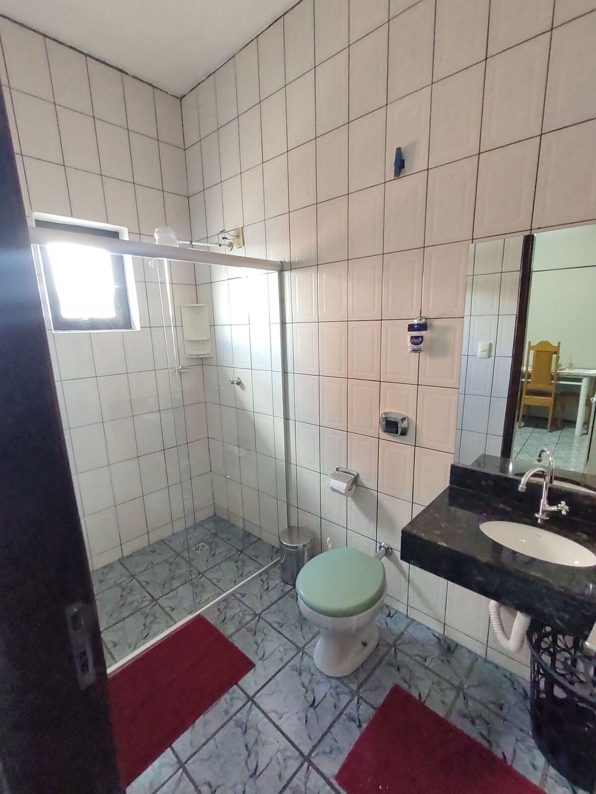 The bathroom features tiled walls and a glass shower enclosure. A white sink with a dark countertop is paired with a mirror. A light green toilet is positioned adjacent to the stall. Two red bath mats are laid on the gray tiled floor.