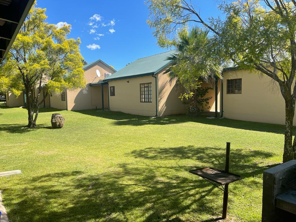A well-maintained outdoor area features a lush green lawn surrounded by trees. Two structures are visible, showcasing a combination of light-colored walls and green roofs. A small barbecue grill is positioned on a concrete slab, providing a space for outdoor cooking.