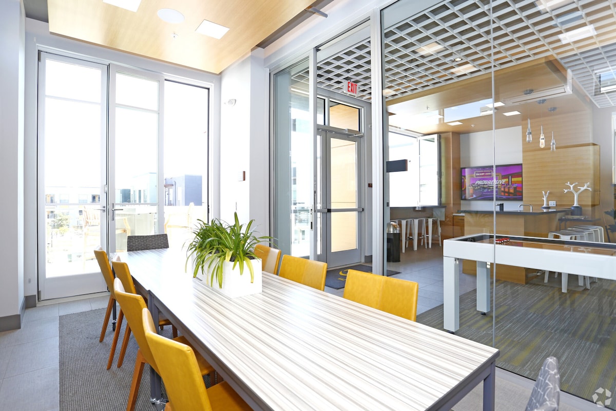 A bright meeting room features a long table surrounded by orange chairs, creating a collaborative atmosphere. Large glass doors allow natural light to fill the space, while modern decor includes minimalistic plants and a nearby presentation area visible through transparent walls.