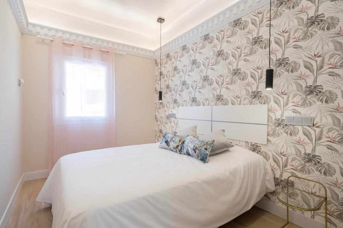 A bright bedroom features a spacious bed adorned with decorative pillows and a light-colored comforter. The wall behind is covered in a floral wallpaper, while a window allows natural light to filter in, complemented by sheer pink curtains. A small side table adds to the decor.