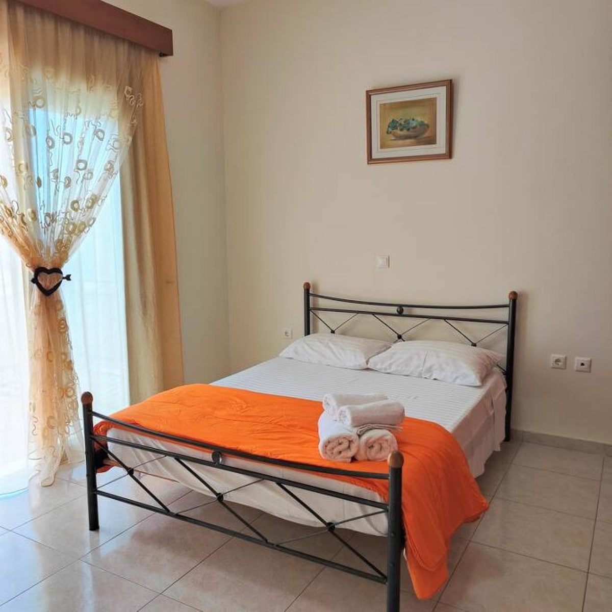 A simple bedroom features a black metal frame bed dressed with a white blanket and an orange throw. Fresh towels are neatly arranged at the foot of the bed. Natural light enters through sheer curtains, and a framed artwork is displayed on the wall.