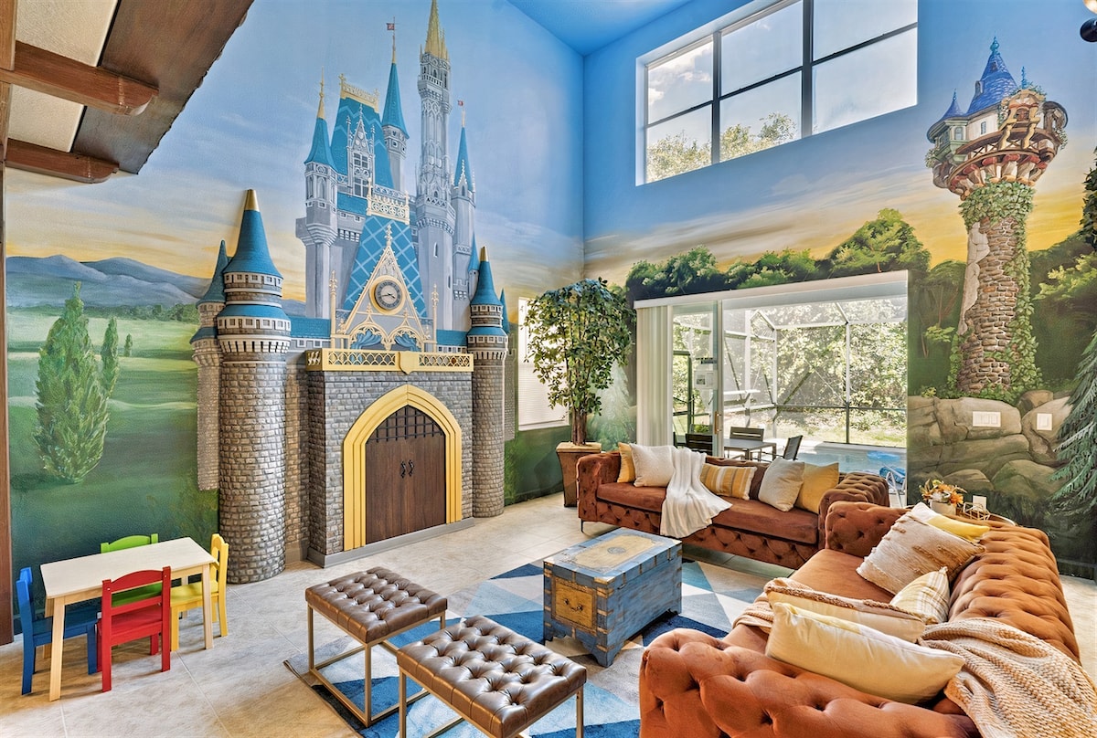 The living room showcases a castle-themed mural featuring a fairy tale castle and a tower. Light floods in through large windows, illuminating the plush brown sofas and colorful rug below. A small play area with a table and chairs is set for children, while a pool view is visible through the sliding glass door.