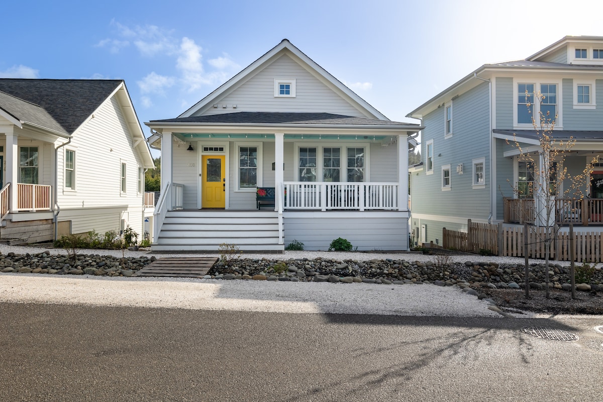 A charming, welcoming home features a light exterior with a bright yellow door and a spacious front porch. Landscape design includes stone pathways and greenery, with neighboring homes visible to the sides, set against a clear blue sky.