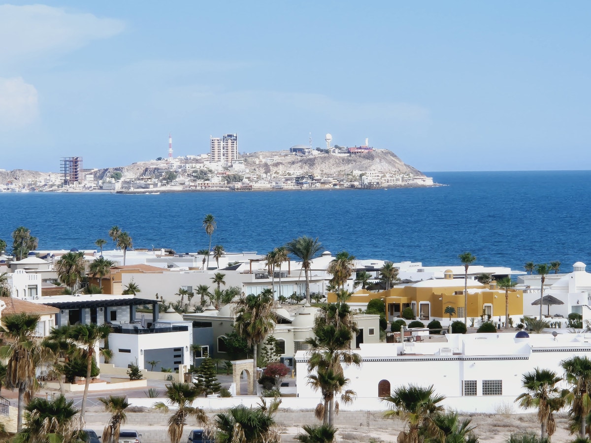 A scenic view showcases the coastline with the ocean in the foreground. Palm trees are seen in the lower part of the image, while distant buildings and structures are positioned along the coastline and on the nearby hills.
