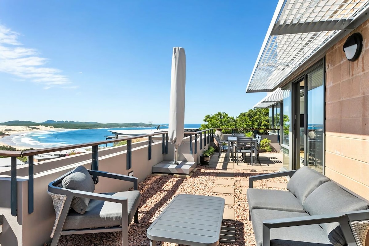 A spacious outdoor terrace overlooks the coastline, featuring comfortable seating arrangements and a dining area. Sunlight bathes the area, where a large umbrella is positioned for shade. Coastal views extend across the horizon, with the beach visible in the distance.