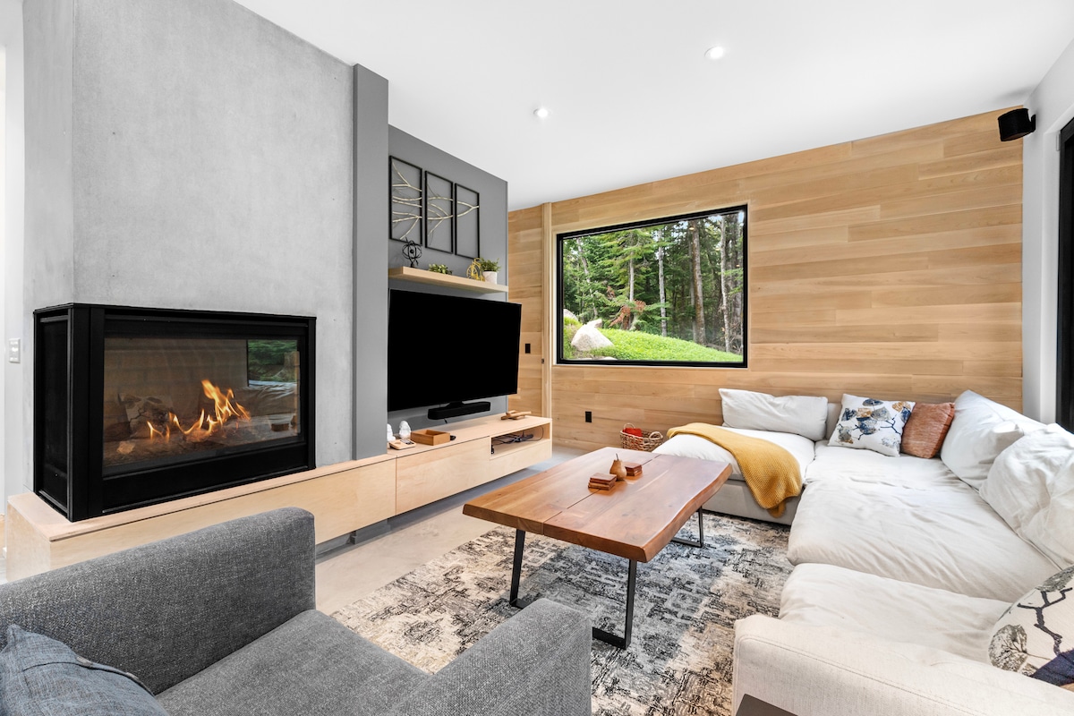 An inviting living area features a modern design with a large sectional sofa and a sleek coffee table. A cozy fireplace adds warmth, while a wide window showcases the lush outdoors, promoting a connection with nature. Soft lighting highlights the natural wood accents.