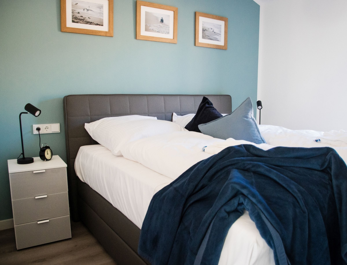 A spacious sleeping area features a large box spring bed with crisp white linens and a plush dark blue throw. A nightstand with a clock lamp stands beside the bed. Light teal walls are adorned with framed beach photographs, creating a calming ambiance.