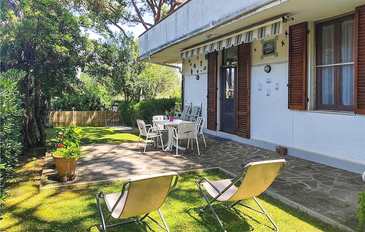 An outdoor patio area features a stone floor, surrounded by greenery. Two lounge chairs are positioned on the grass, with a dining table and chairs set under a covered area. Decorative plants and a wooden fence enhance the inviting atmosphere.