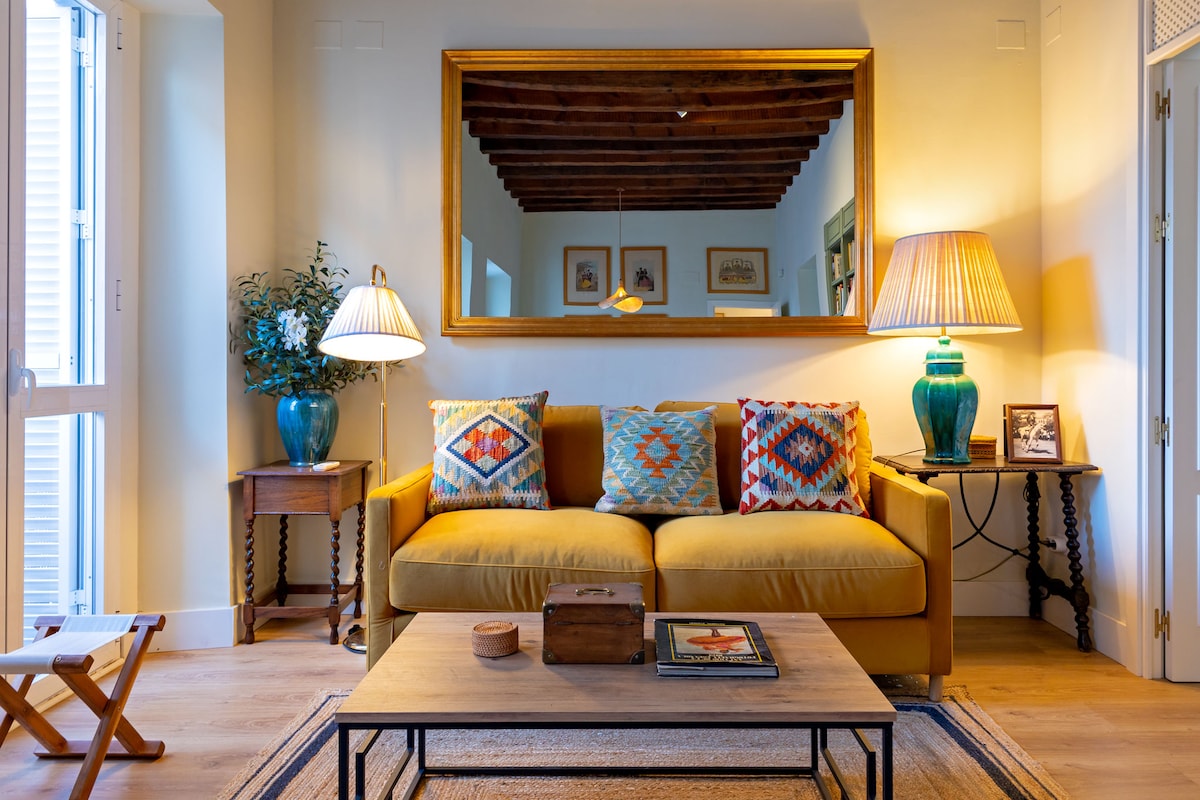 A cozy living area is featured with a yellow sofa adorned with colorful cushions. A large mirror reflects the room's light, complemented by two stylish lamps. A wooden coffee table is placed centrally, with decorative items visible on its surface, enhancing the flat's inviting atmosphere.