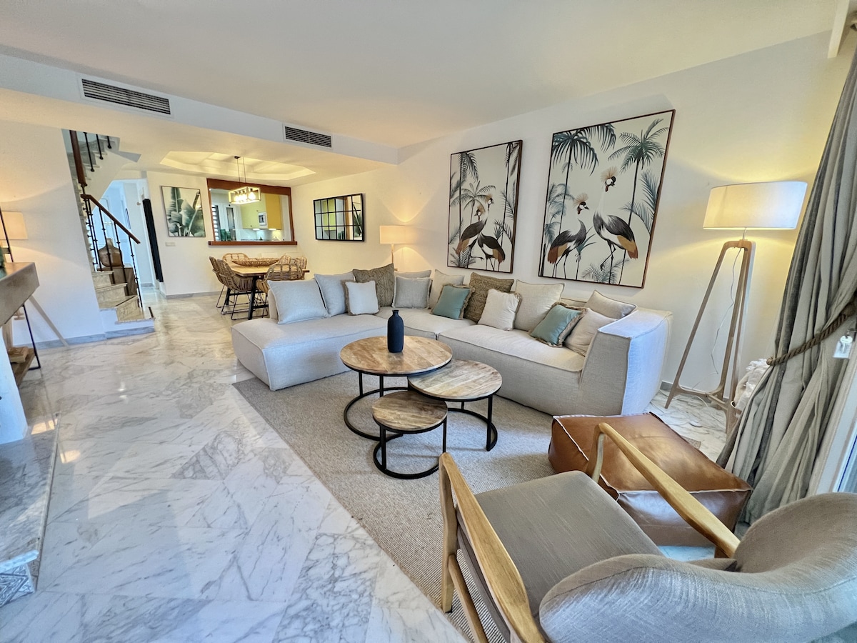 A spacious living area features a light-colored sofa adorned with decorative pillows, surrounded by wooden coffee tables. Large artwork depicting tropical scenes is displayed on the wall. Natural light filters through large windows, enhancing the elegant marble flooring.
