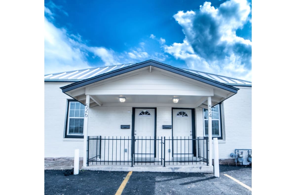 The exterior of a well-maintained building features a covered entrance with a peaked roof. Two doorways are framed by windows on either side, while a black railing adds a touch of elegance. The expansive blue sky above is complemented by a few clouds, enhancing the inviting look.