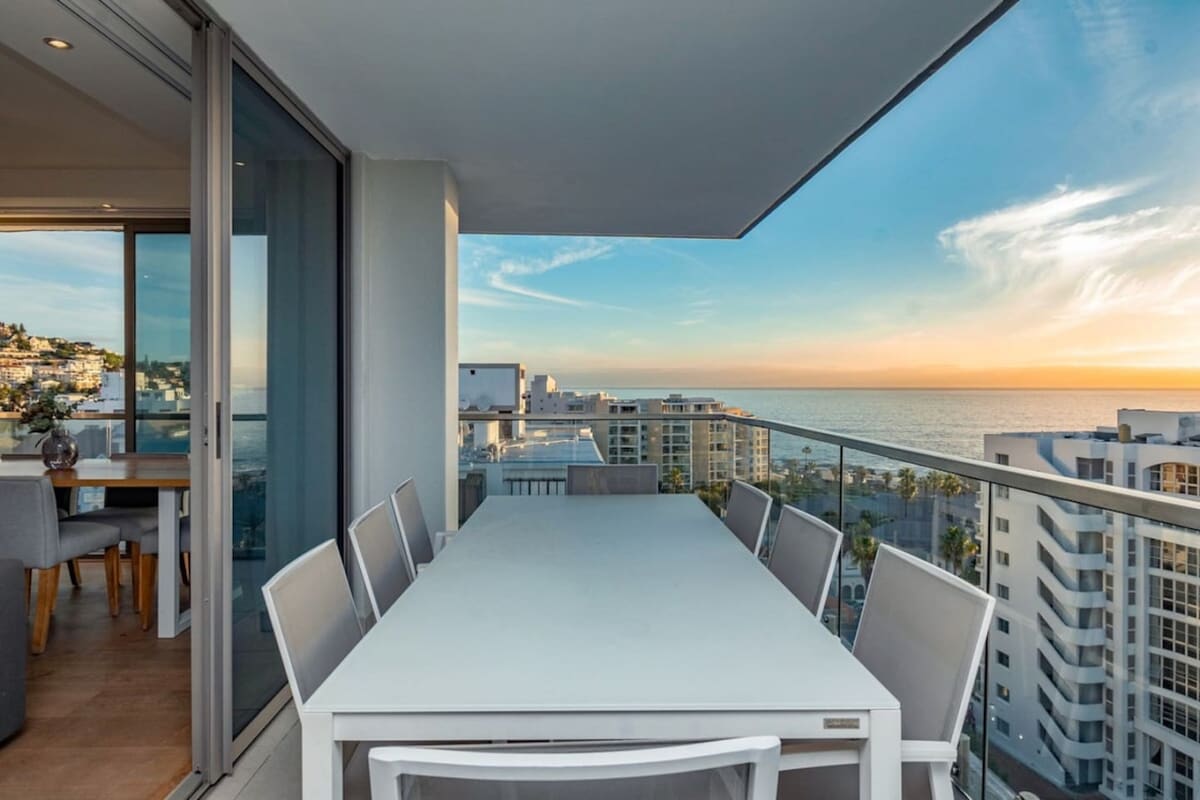 An outdoor dining area is presented on the balcony, featuring a large white table surrounded by multiple chairs. The ocean and sunset can be seen in the background, with soft colors filling the sky. Large glass doors lead back into the living area.