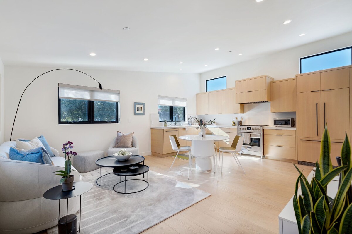 An open-plan living area is presented, featuring modern wooden cabinetry and sleek appliances. A white dining table is surrounded by four chairs, while a comfortable sofa sits beside a coffee table on a light rug. Large windows allow natural light to fill the space.