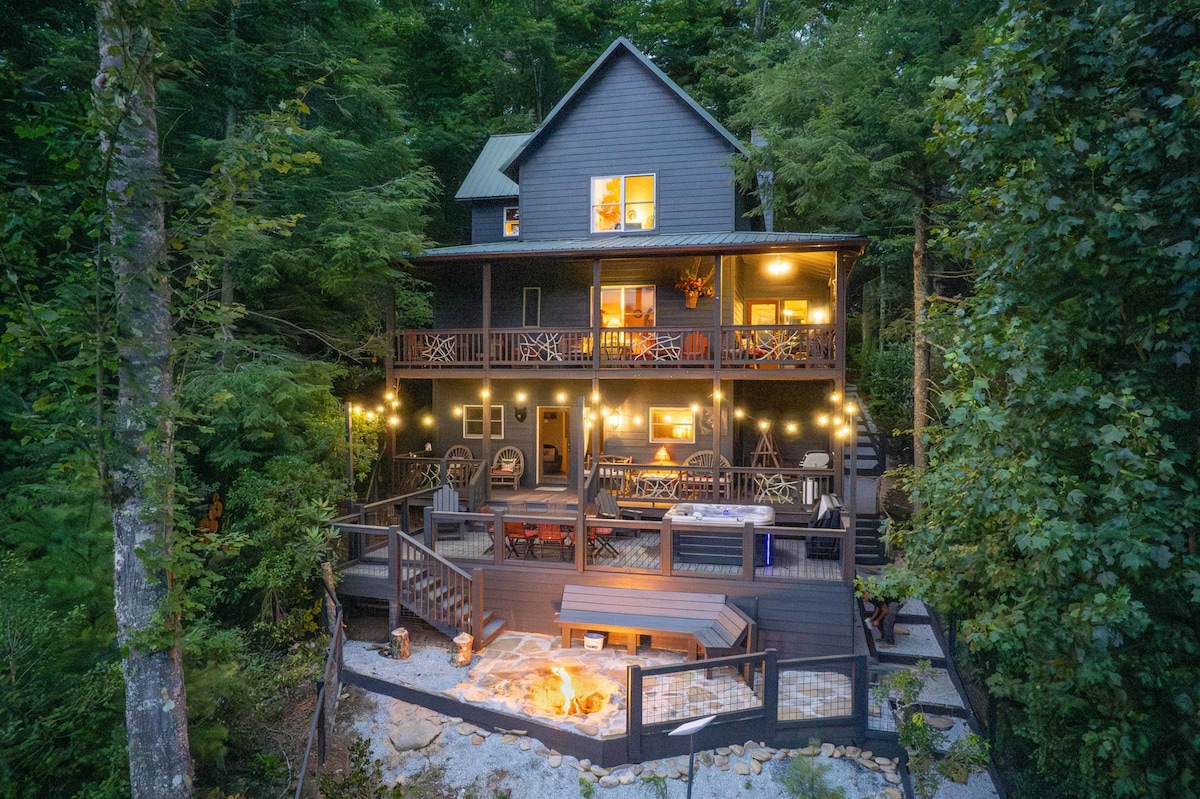 The cabin showcases a multi-level structure surrounded by tall trees, illuminated by warm lights. Various seating areas, including rocking chairs and a firepit, are visible on the deck. A hot tub is nestled on the lower level, offering a serene outdoor retreat.