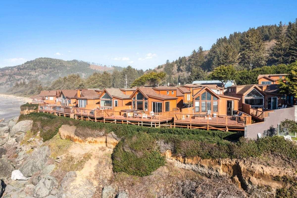 A row of charming oceanfront cabins is positioned along a natural cliff, surrounded by coastal vegetation. The buildings feature wooden exteriors and large windows, capturing views of the Pacific Ocean. The landscape includes a sandy beach and a backdrop of gentle hills under clear blue skies.