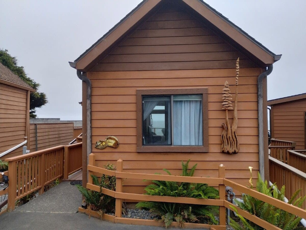 The exterior of the cabin features warm wooden siding and a welcoming entrance. A window offers a glimpse of the interior, while a decorative piece resembling ocean life is mounted on the wall. A well-maintained pathway leads to the entrance, surrounded by greenery.