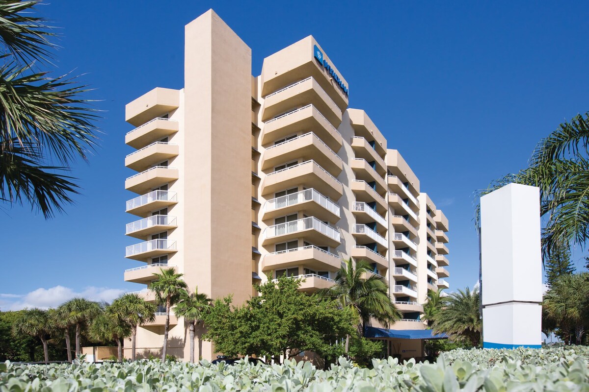A modern resort building is prominently displayed against a clear blue sky. The structure has multiple balconies with light beige walls, flanked by lush greenery and palm trees. The ground is covered with low shrubs, enhancing the inviting atmosphere of the property.