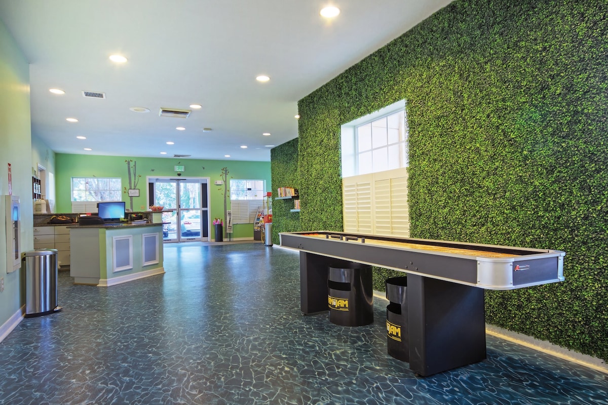 The activities center features a vibrant green wall and spacious layout. A shuffleboard table is positioned in the foreground, creating a game area. Natural light streams in through large windows, illuminating the room and enhancing the inviting atmosphere with a modern design.