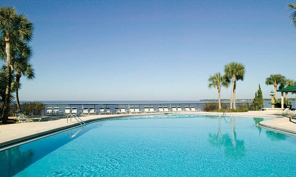 An outdoor swimming pool is surrounded by lounge chairs, with palm trees lining the perimeter. The clear blue water reflects the bright sky, and the calming view extends towards the water beyond the pool area.