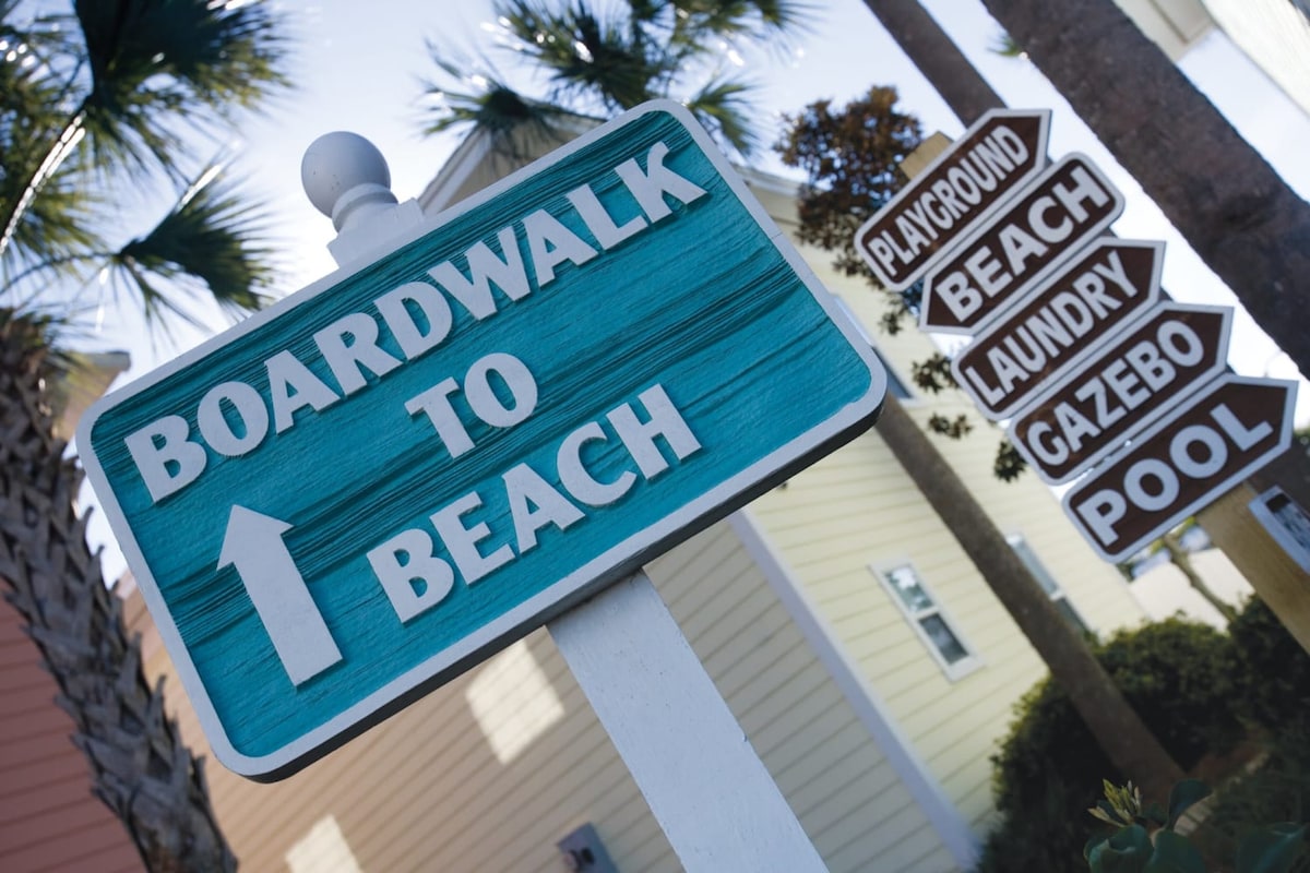 A signpost features directional indicators to various amenities, including the beach, pool, playground, gazebo, and laundry facilities. The vibrant turquoise sign for 'Boardwalk to Beach' stands prominently, framed by palm trees and colorful buildings in the background.