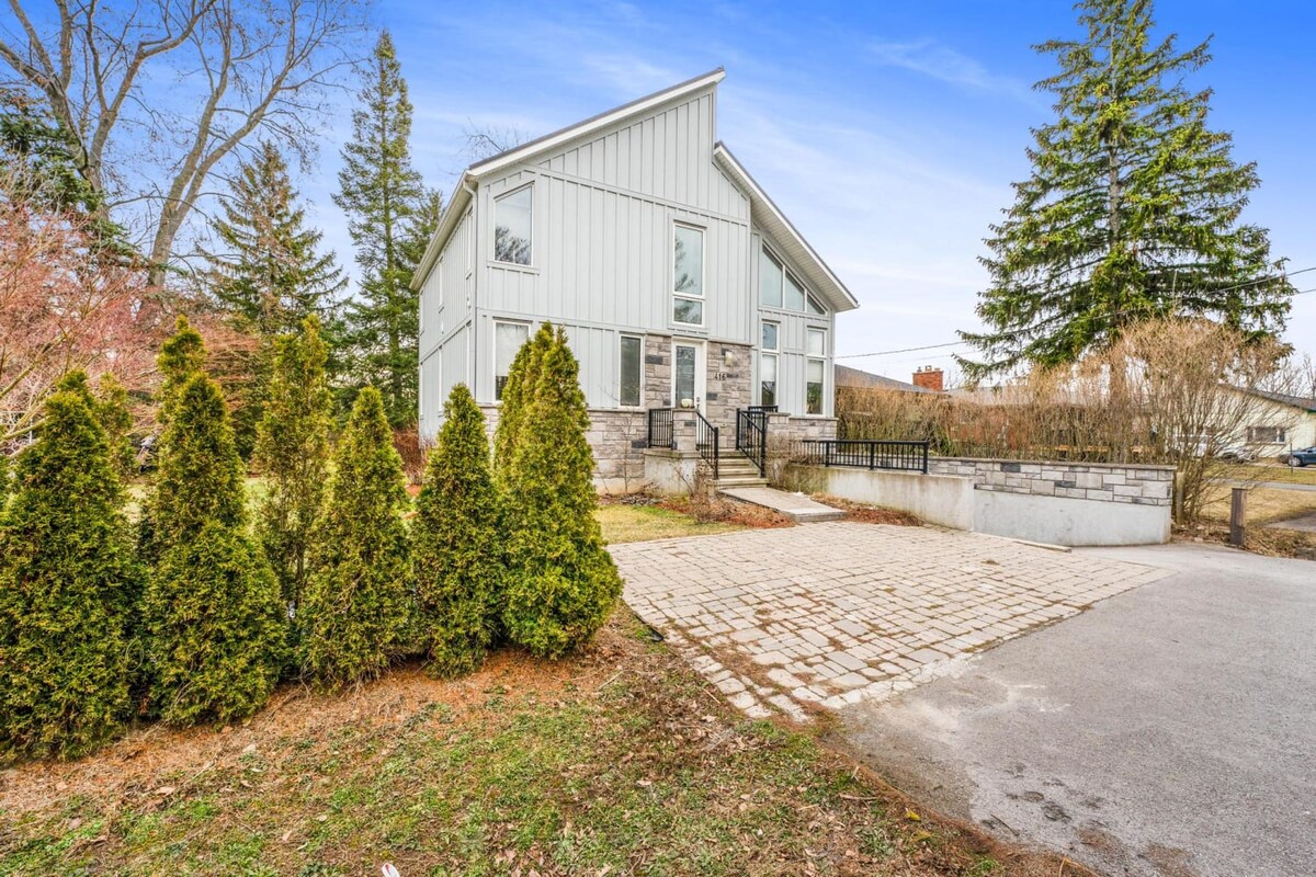 A modern two-story house is depicted with a light-colored exterior. A stone pathway leads to the entrance, framed by neatly manicured hedges. Large windows allow natural light to fill the space, while surrounding trees add to the inviting outdoor ambiance.