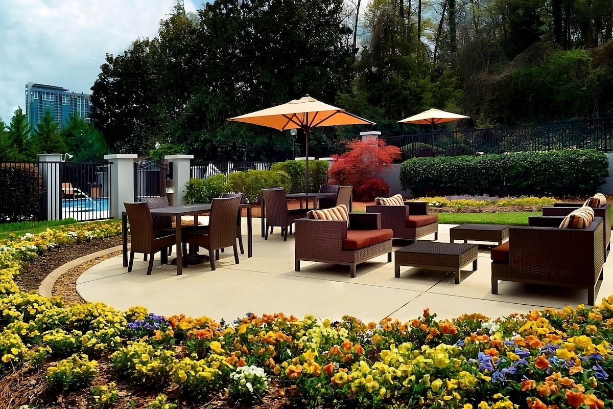 An outdoor relaxation area is surrounded by a vibrant flower bed, featuring comfortable seating and a dining table shaded by large umbrellas. The scene is complemented by lush greenery and a glimpse of the swimming pool in the background.