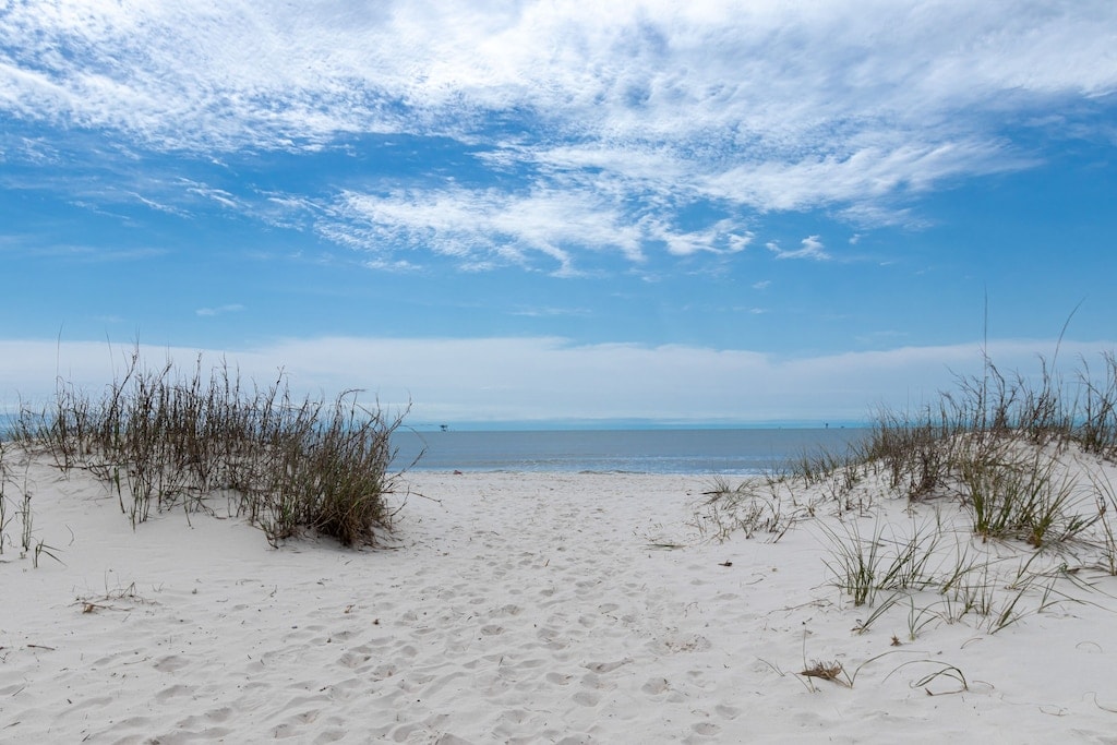 A sandy pathway leads from the beach grass to the water, with gentle waves lapping at the shoreline. The sky is filled with soft clouds, creating a serene atmosphere. The tranquil view showcases the expansive Gulf of Mexico under bright blue hues.