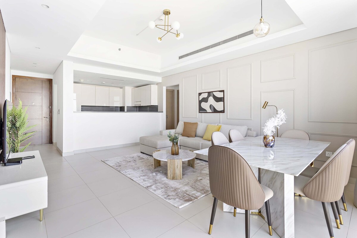The living and dining area offers a spacious layout, featuring a large sectional sofa and a stylish round dining table with four chairs. A decorative rug adds warmth to the space, while modern artwork decorates the wall, complemented by natural light flowing in through wide windows.