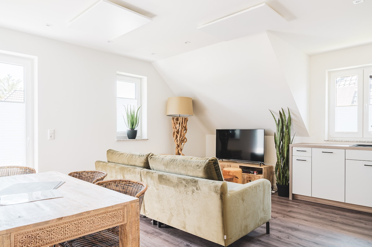An airy living space features a light-colored couch positioned between two windows. A dining area with a rustic wooden table complements the room, while a modern television sits on a wooden stand adjacent to a green plant. The overall design is minimal and functional.