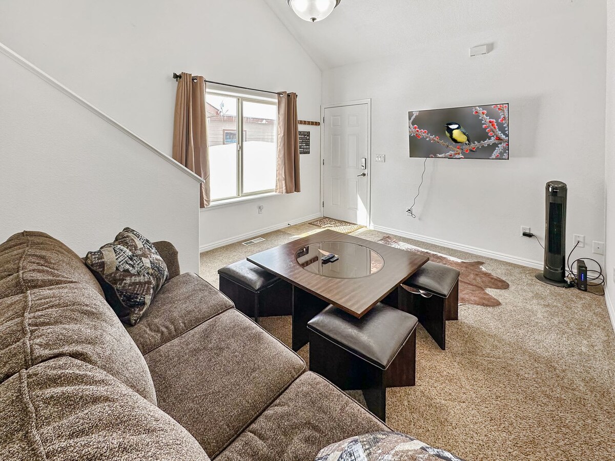 A comfortably arranged living area is highlighted by a spacious sectional sofa. A low table with seating is centered in the room, complemented by a large window allowing natural light to fill the space. A wall-mounted television adds to the inviting atmosphere.
