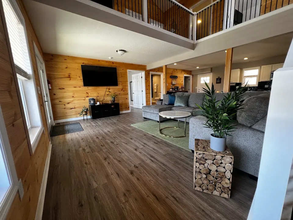 An open living space features a spacious gray sectional sofa and a circular coffee table centered on a soft rug. Natural wood walls and large windows allow ample light. A flat-screen TV is mounted on the wall, providing entertainment options.