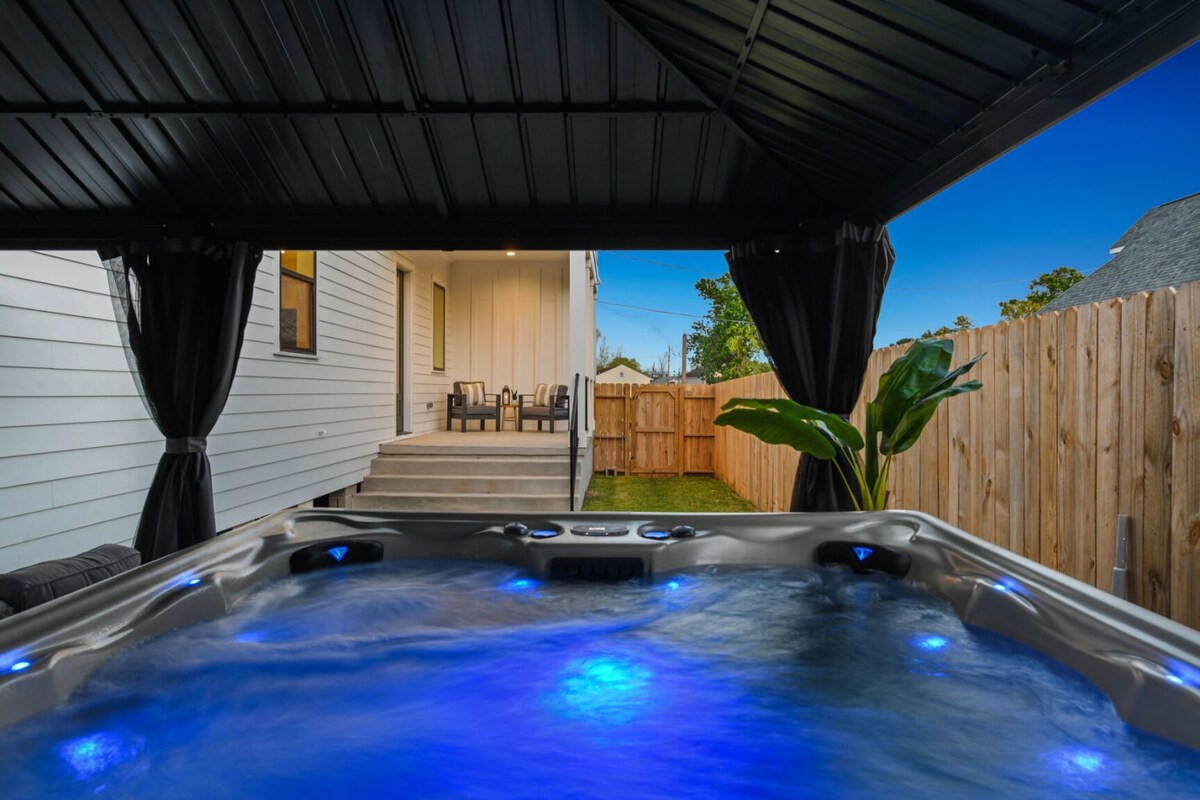 A hot tub is positioned under a gazebo, with blue lights illuminating the water. The background reveals a backyard space featuring steps leading to a deck with outdoor seating. The surrounding fence and plants add to the cozy ambiance.