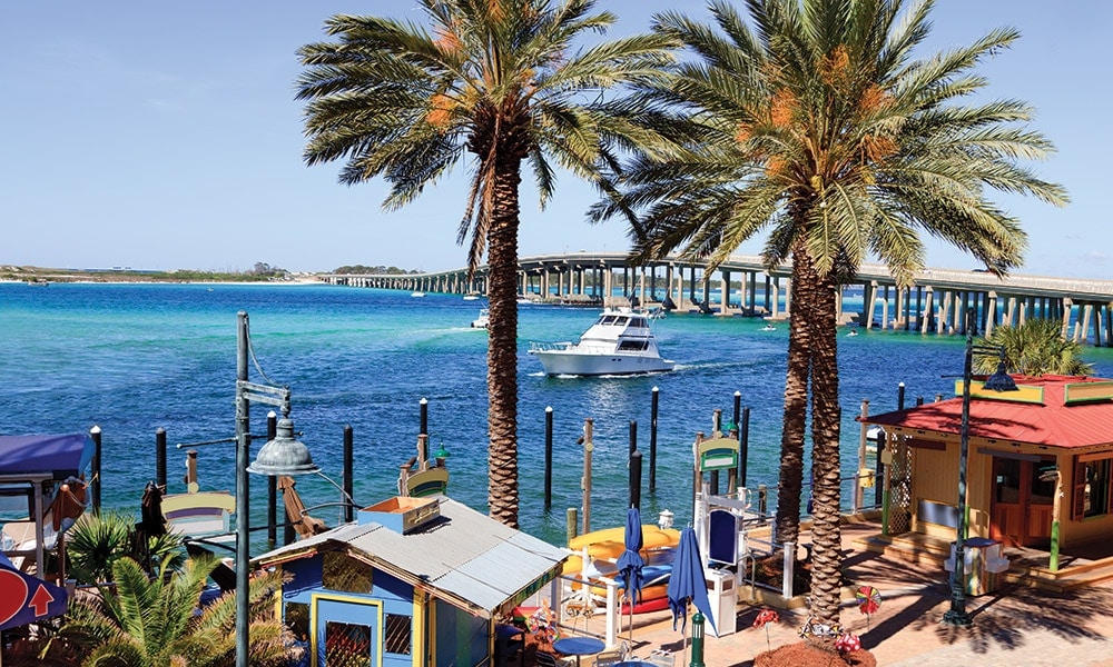 A scenic view of the waterfront showcases vibrant blue waters and a distant bridge. Palm trees provide shade over colorful structures, while a boat glides gracefully across the water, emphasizing the lively atmosphere of the area.