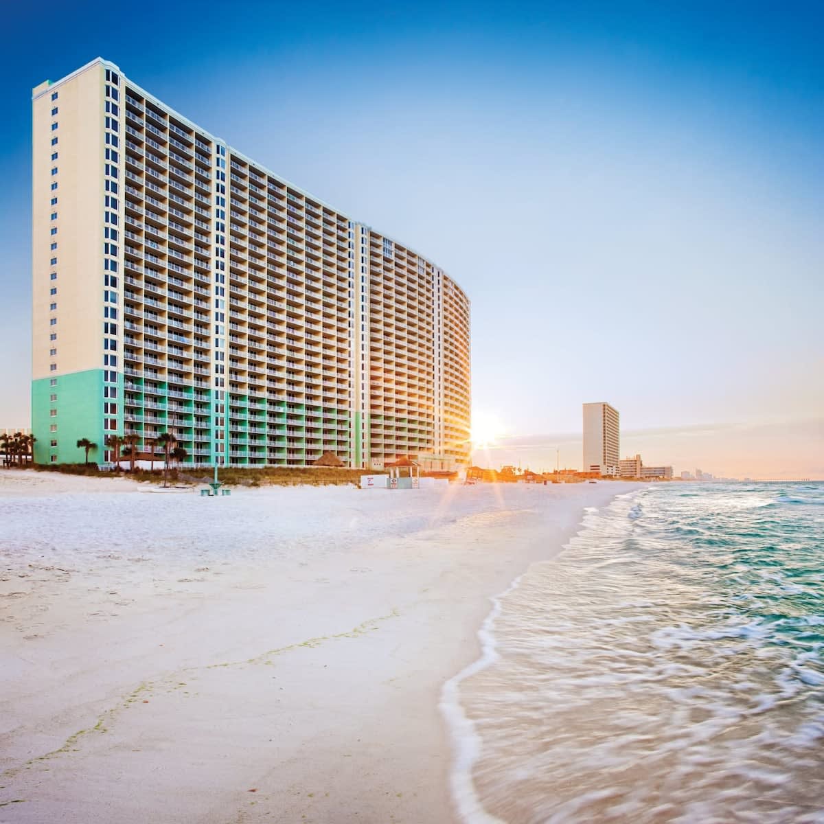 A tall resort building curves gracefully along the shoreline, with its façade reflecting hues of light blue and turquoise. Gentle waves lap against the sandy beach, shimmering in the warm sunlight, while the sunrise casts a soft glow across the horizon.