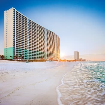 A tall, contemporary beachfront building stands along a sandy shore, reflecting the colors of the sunset. Gentle waves lap at the beach, creating a serene atmosphere. The coastline stretches in the background, showcasing the expansive sky above.