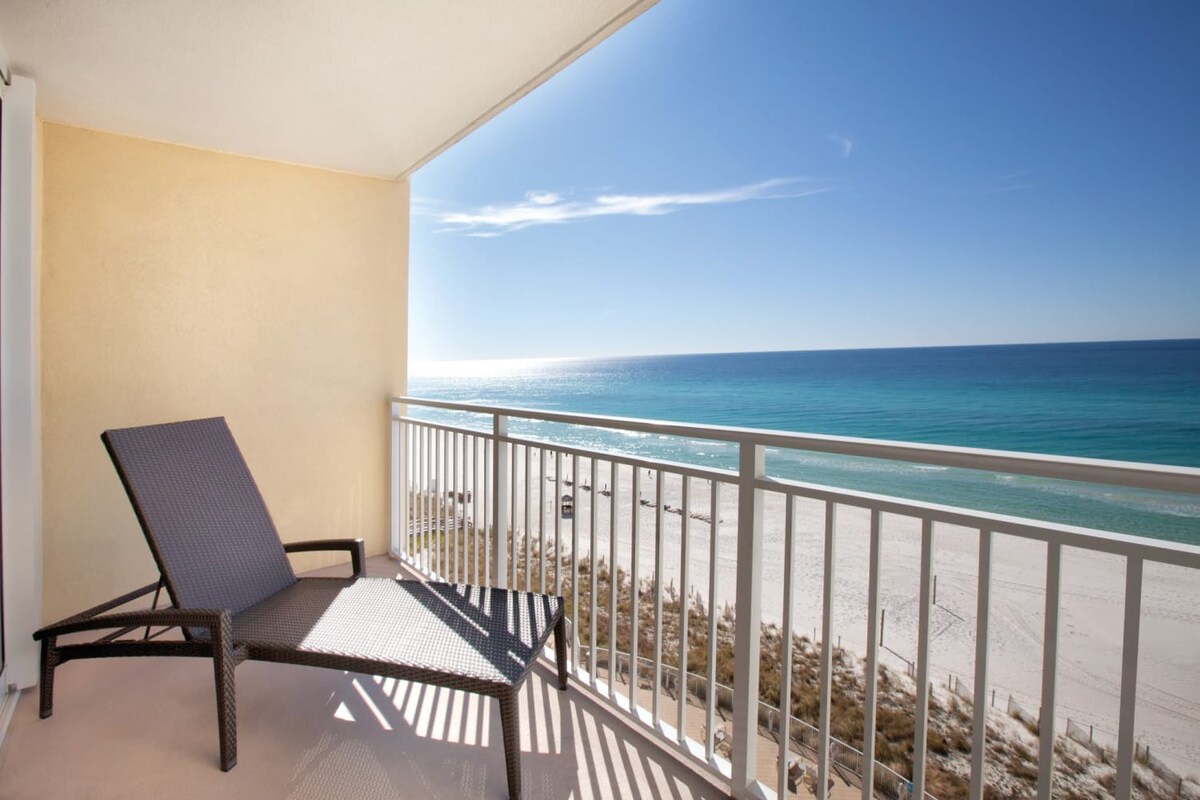 A shaded balcony features a single lounge chair facing the sparkling waters of the Gulf. The expansive view reveals soft sandy beaches and clear blue skies, inviting relaxation and enjoyment of the coastal scenery.