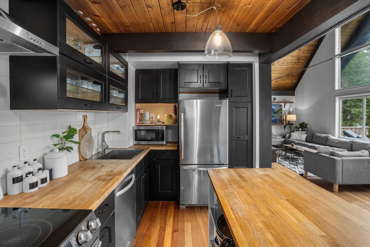 A modern kitchen is highlighted by sleek black cabinetry and stainless steel appliances. Warm wood countertops provide ample workspace. Shelving displays glassware, while decorative plants add a touch of greenery. Natural light enters through a nearby window, illuminating the kitchen's functional design.