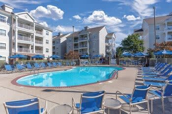 An outdoor swimming pool is surrounded by several lounge chairs, all neatly arranged. Umbrellas provide shade nearby, while the three-story building serves as backdrop against a bright blue sky dotted with fluffy white clouds.