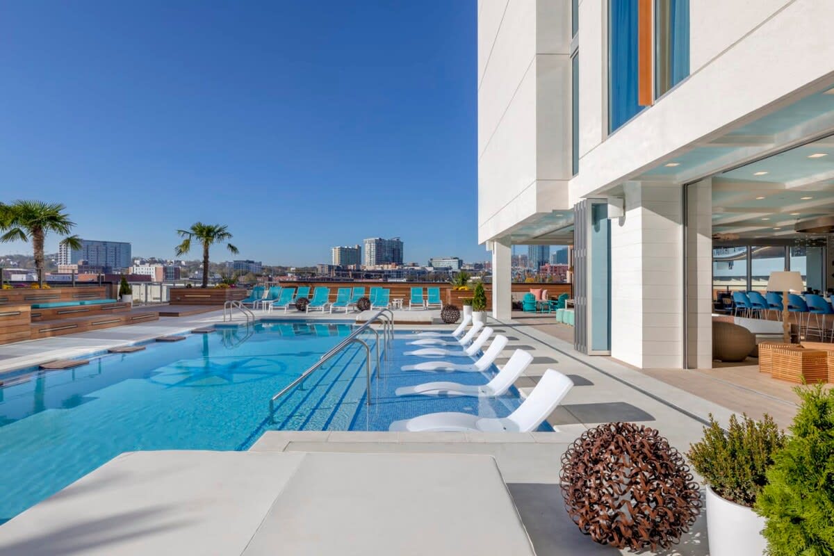 The outdoor pool area features sun loungers arranged around a clear blue pool. Palm trees provide shade, while modern architectural lines and seating areas extend around the pool, offering panoramic views of the city skyline in the distance.