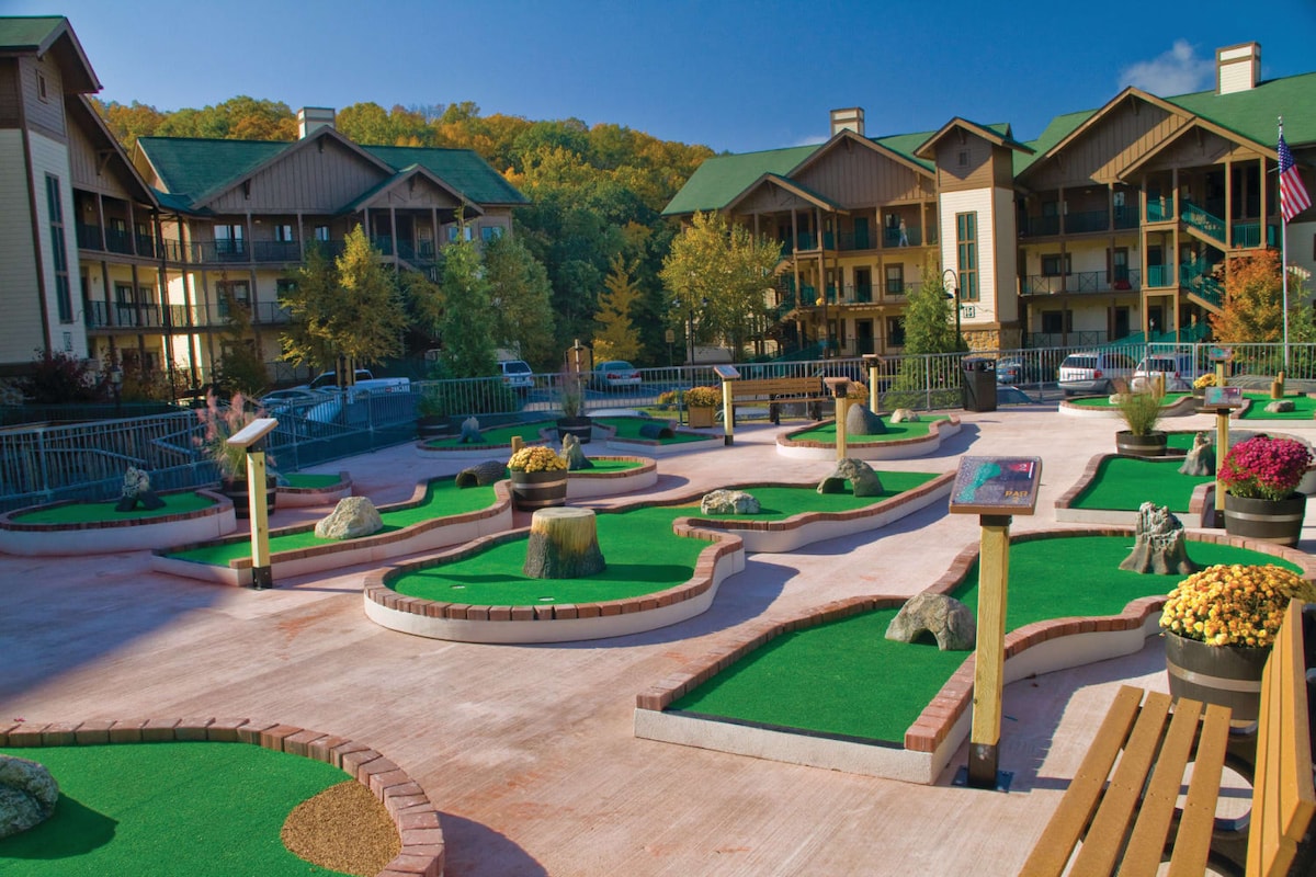 An outdoor miniature golf course is depicted, featuring green carpets, sandy areas, decorative rocks, and flower planters. Surrounding buildings with a mix of wood and stone architecture are visible in the background, along with vibrant autumn foliage on the distant hills.