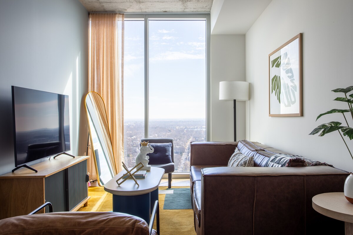 A modern living area displays a spacious sofa beside a large window offering expansive views. A flat-screen television sits on a wooden console, with a standing mirror and a comfortable chair positioned nearby. Natural light fills the room, creating an inviting space.