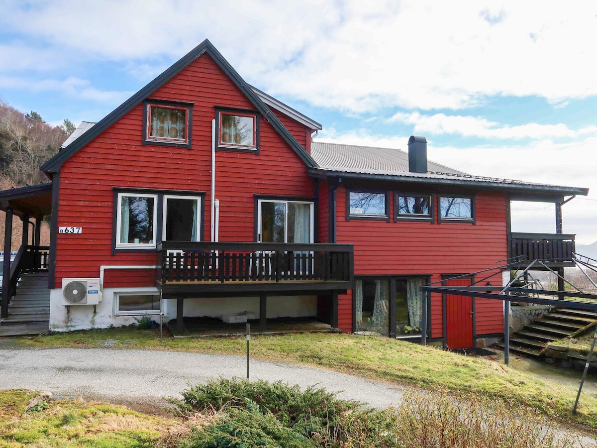 A red two-story house features a sloped roof and multiple windows on both levels. A black balcony extends from the upper floor, while a staircase leads to the main entrance. Surrounding greenery adds to the property’s natural setting.