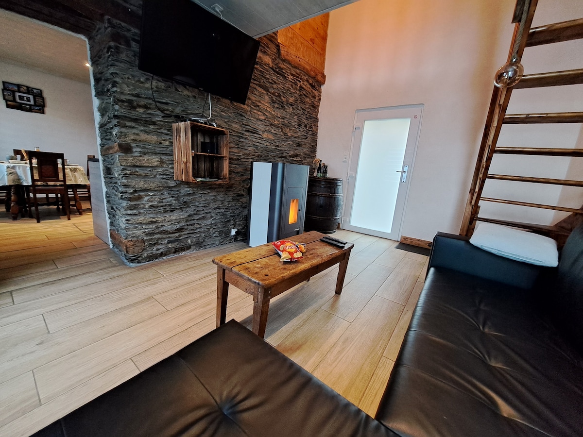 A cozy living area features a black sofa positioned beside a rustic wooden coffee table. A flat-screen TV is mounted on the stone wall. Natural light enters through the door, leading outside, while a warm glow is emitted from a free-standing heater.