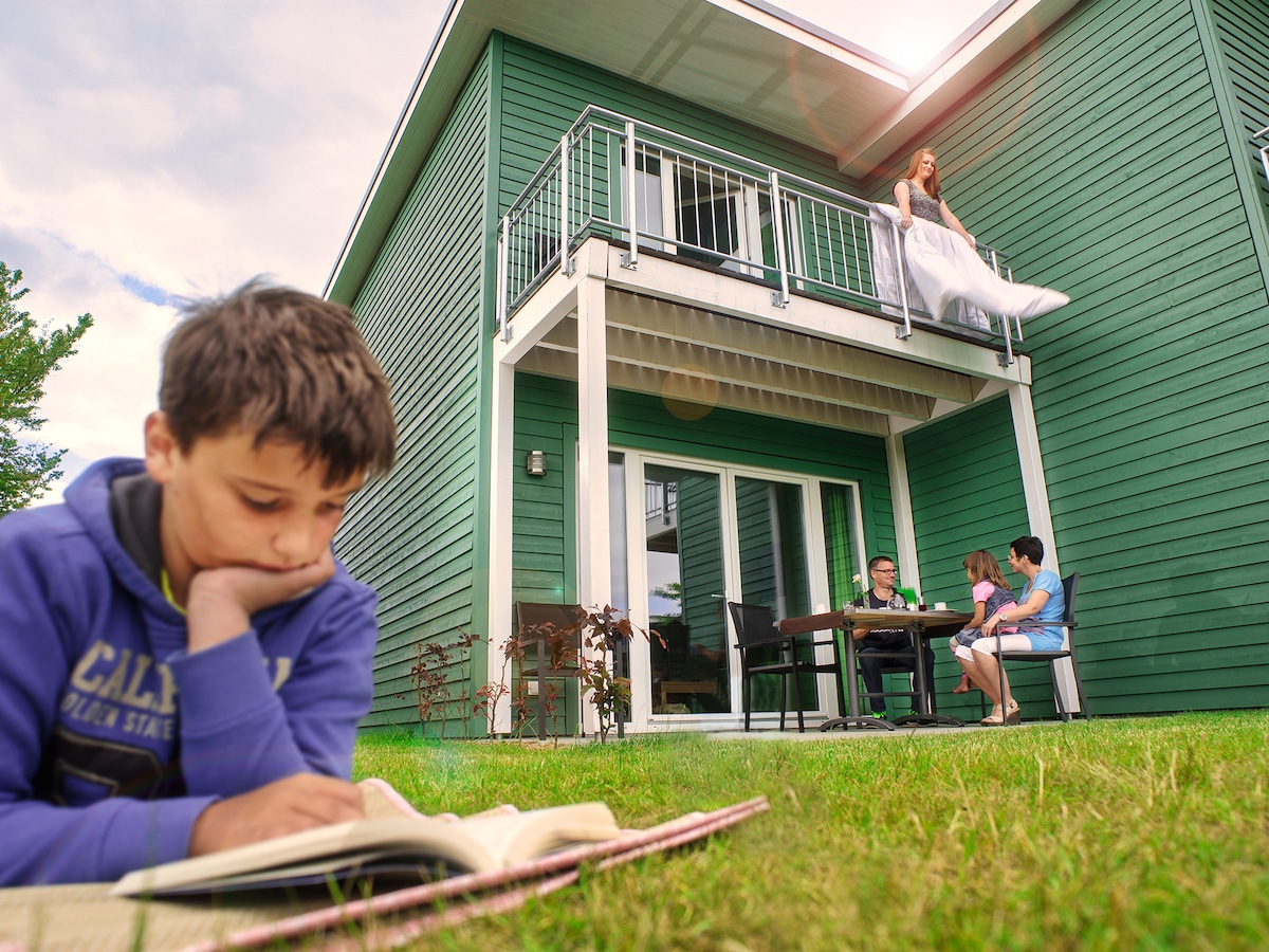 A green, modern building is shown with a balcony on the second floor. A child reads on the grass, while a family gathers around a dining table outside. The scene captures a lively and relaxed outdoor atmosphere.