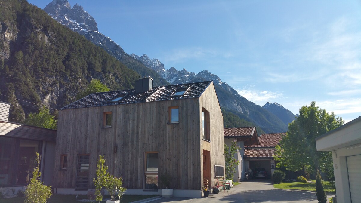A modern wooden house is set against a backdrop of majestic mountains under a clear blue sky. The structure features large windows and a pitched roof, while the surrounding area includes greenery and parked vehicles, contributing to the serene landscape.