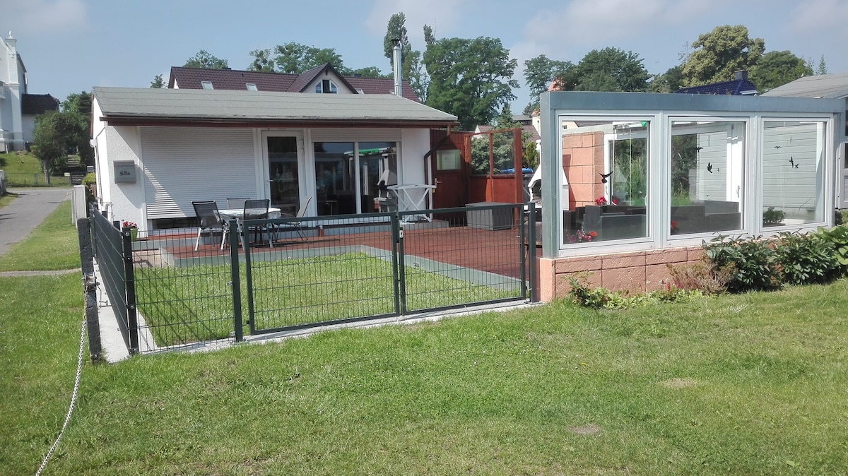 A single-story bungalow is surrounded by a fenced yard, featuring a patio with outdoor seating. The exterior showcases a combination of stone and glass elements, highlighting an inviting entrance. Lush greenery and trees provide a calm backdrop, under a clear blue sky.