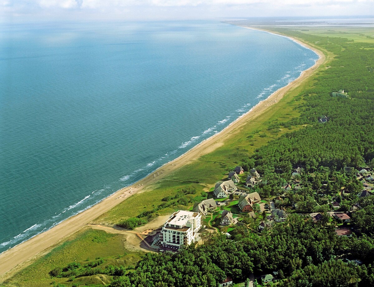 An aerial view captures a serene coastline, where soft sandy beaches meet the calm blue ocean. Lush greenery lines the shore, with a hotel structure visible among coastal residences. The expansive sky complements the tranquil waters, showcasing a peaceful retreat by the sea.