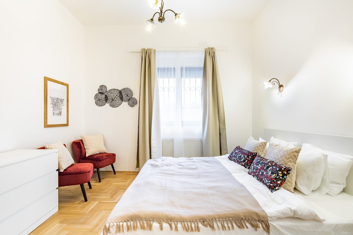 A bright bedroom features a spacious bed adorned with decorative pillows and a woven throw. Two red armchairs provide a cozy seating area beside a white dresser. Natural light streams through a window, complemented by elegant curtains that frame the view.