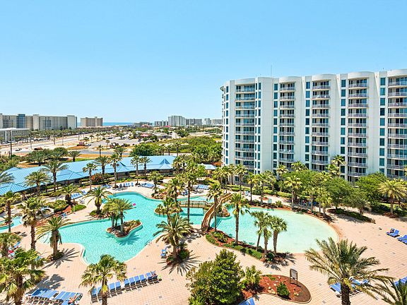 A scenic view of a resort's expansive lagoon pool is depicted, surrounded by lush palm trees and tropical landscaping. Multiple loungers are arranged around the pool area, and the nearby buildings reflect the vibrant colors of the surroundings. The ocean is visible in the distance.